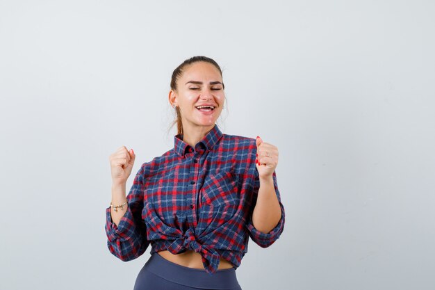 Mujer joven en camisa a cuadros, pantalones mostrando gesto de ganador y mirando dichoso, vista frontal.