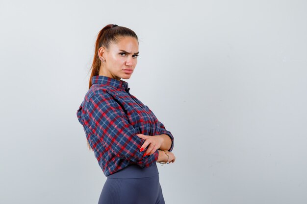 Mujer joven en camisa a cuadros, pantalones de pie con los brazos cruzados y mirando confiado, vista frontal.