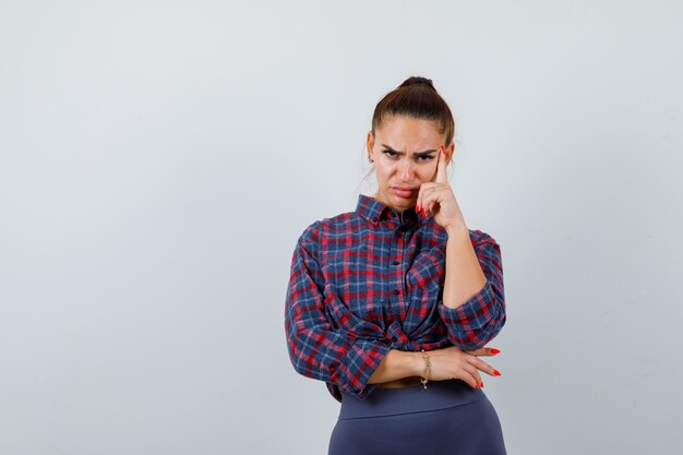 Mujer joven en camisa a cuadros, pantalones de pie en pose de pensamiento y mirando pensativo, vista frontal.