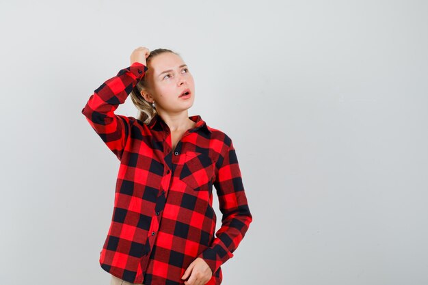 Mujer joven en camisa a cuadros, pantalones rascándose la cabeza, mirando hacia arriba y mirando vacilante, vista frontal.