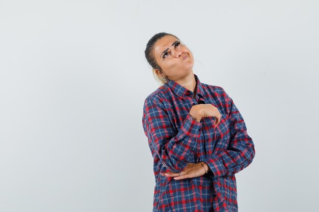 Mujer joven en camisa a cuadros de pie en pose de pensamiento y mirando pensativo, vista frontal.