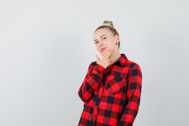 Mujer joven en camisa a cuadros posando con los dedos tocando la barbilla y luciendo bonita