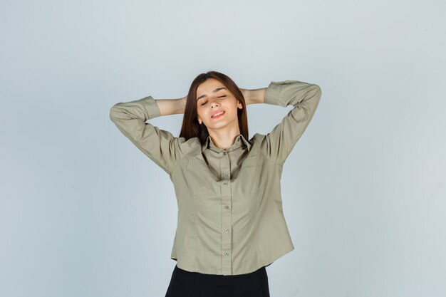 Foto gratuita mujer joven en camisa, falda manteniendo las manos detrás de la cabeza y mirando relajado, vista frontal.