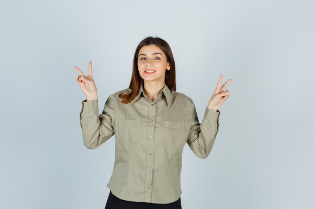 Mujer joven en camisa, falda mostrando gesto de paz y mirando alegre, vista frontal.