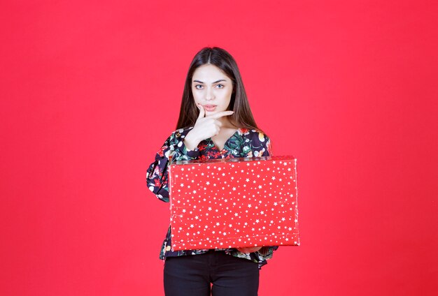 Mujer joven en camisa floral sosteniendo una caja de regalo roja con puntos blancos y parece confundida y pensativa