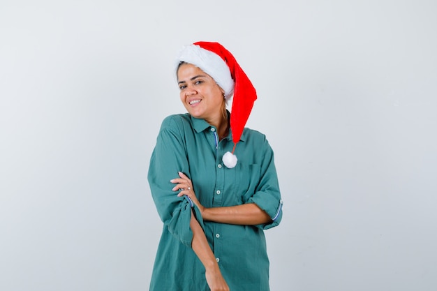 Mujer joven en camisa, gorro de Papá Noel con la mano en el brazo mientras sonríe y mira alegre, vista frontal.