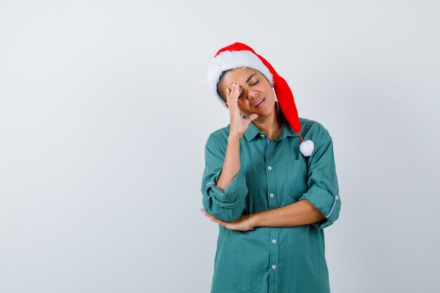 Mujer joven en camisa, gorro de Papá Noel manteniendo la mano en la cabeza y con aspecto cansado, vista frontal.