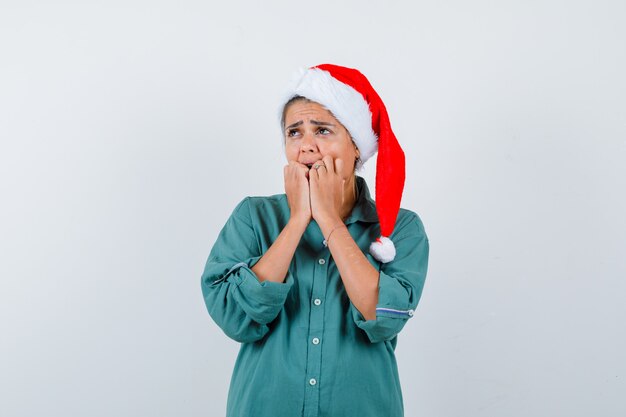 Mujer joven en camisa, gorro de Papá Noel manteniendo las manos en la cara y mirando horrorizado, vista frontal.