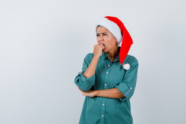 Mujer joven en camisa, gorro de Papá Noel mordiendo el puño emocionalmente mientras mira hacia otro lado y mira ansiosa, vista frontal.