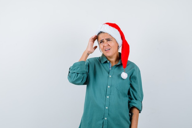 Mujer joven en camisa, gorro de Papá Noel que sufre de dolor de cabeza y luce doloroso, vista frontal.