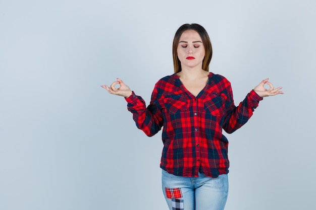 Foto gratuita mujer joven en camisa, jeans mostrando el signo de mudra mientras mantiene los ojos cerrados y parece relajado, vista frontal.