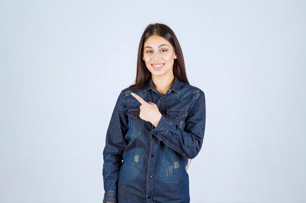 Mujer joven en una camisa de mezclilla apuntando hacia el lado izquierdo con emociones de cara