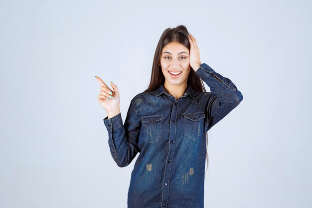 Mujer joven en una camisa de mezclilla apuntando hacia el lado izquierdo con emociones de cara