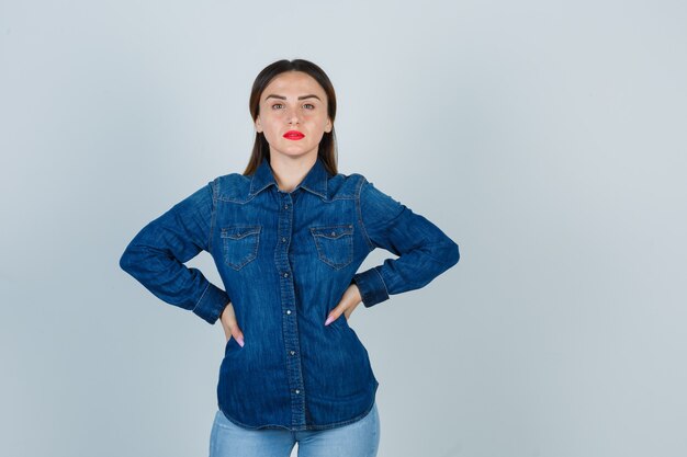 Mujer joven en camisa de mezclilla y jeans tomados de la mano en la cadera y mirando atractivo