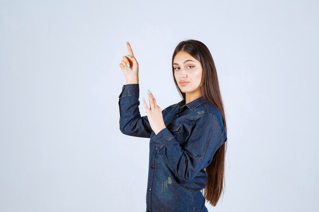 Mujer joven en camisa de mezclilla levantando sus manos y apuntando a algo arriba
