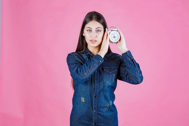 Foto gratuita mujer joven en camisa de mezclilla sosteniendo el despertador en su oído y escuchando