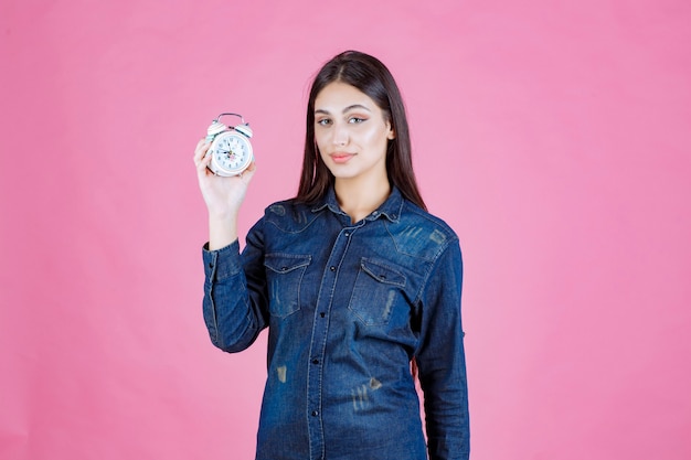 Mujer joven en camisa de mezclilla sosteniendo y promocionando un despertador