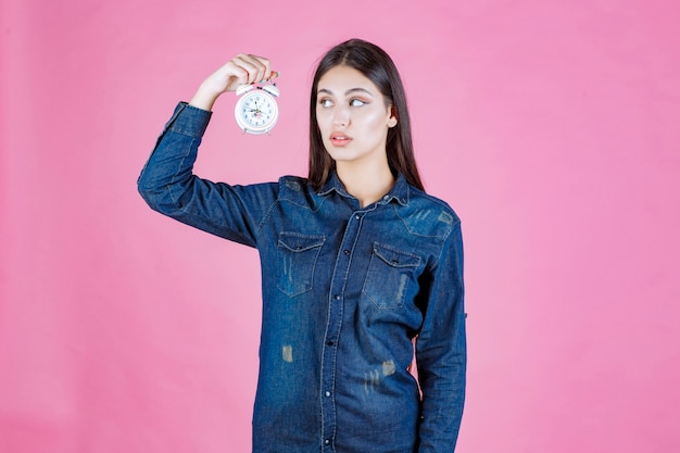 Mujer joven en camisa de mezclilla sosteniendo y promocionando un despertador