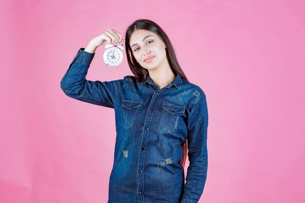 Mujer joven en camisa de mezclilla sosteniendo y promocionando un despertador