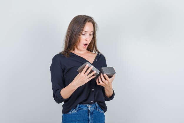 Mujer joven en camisa negra, pantalones cortos de jeans mirando en caja de reloj abierta y mirando sorprendido