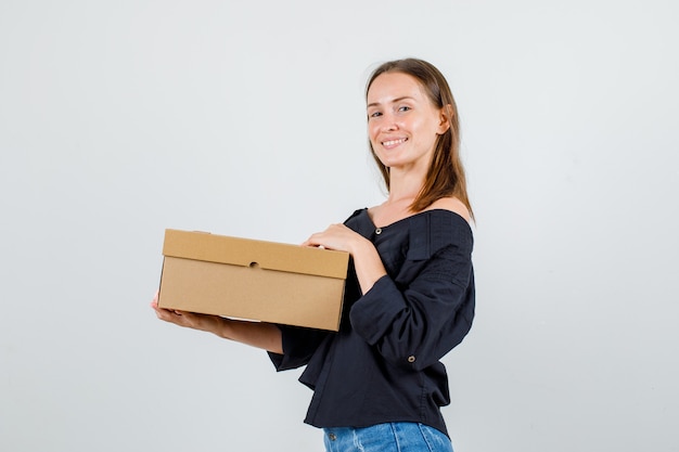 Mujer joven en camisa, pantalones cortos con caja de cartón y sonriendo.