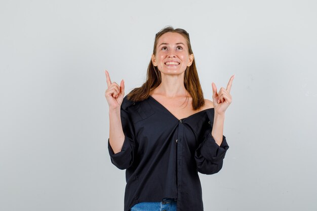 Mujer joven en camisa, pantalones cortos, gafas apuntando hacia arriba con gesto de pistola y mirando alegre