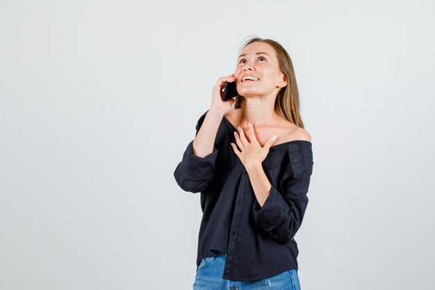 Mujer joven en camisa, pantalones cortos hablando por teléfono inteligente mientras mira hacia arriba y se ve alegre