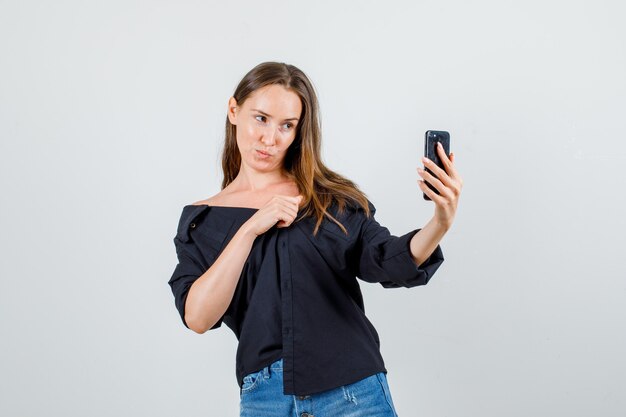 Mujer joven en camisa, pantalones cortos posando mientras toma selfie en teléfono inteligente y se ve linda