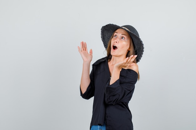Mujer joven en camisa, pantalones cortos, sombrero levantando las manos y mirando sorprendido
