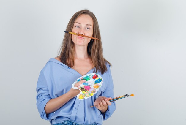 Mujer joven en camisa, pantalones cortos sosteniendo pincel y paleta y mirando divertido