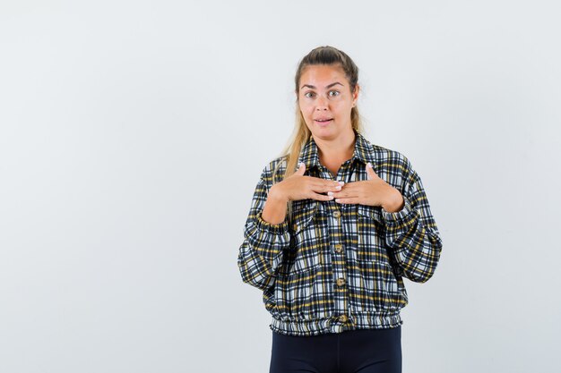 Mujer joven en camisa, pantalones cortos tomados de la mano en el pecho y mirando asombrado, vista frontal.