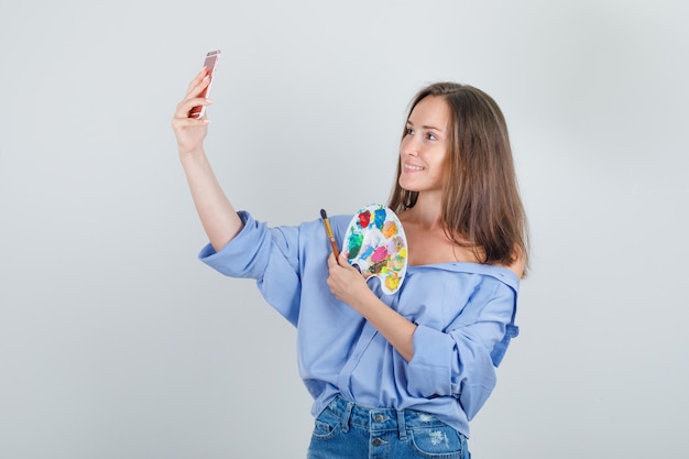 Mujer joven en camisa, pantalones cortos tomando selfie con herramientas de pintura y mirando alegre.