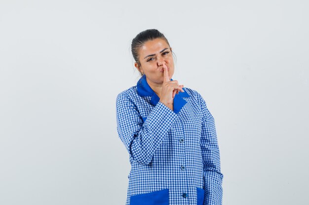 Mujer joven en camisa de pijama de cuadros azules poniendo el dedo índice en el labio, mostrando gesto de silencio y luciendo bonita, vista frontal.