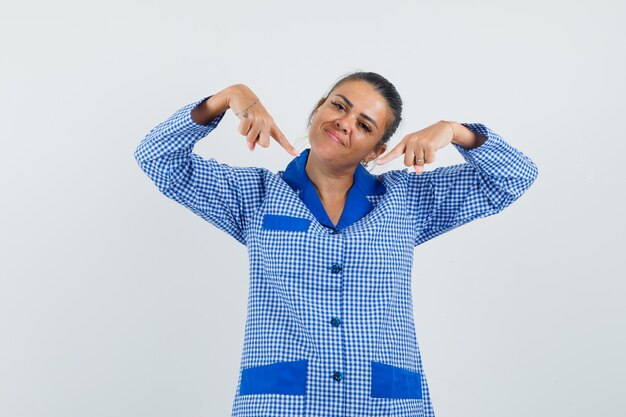 Mujer joven en camisa de pijama de cuadros vichy azul apuntando a sí misma con los dedos índices y mirando feliz, vista frontal.