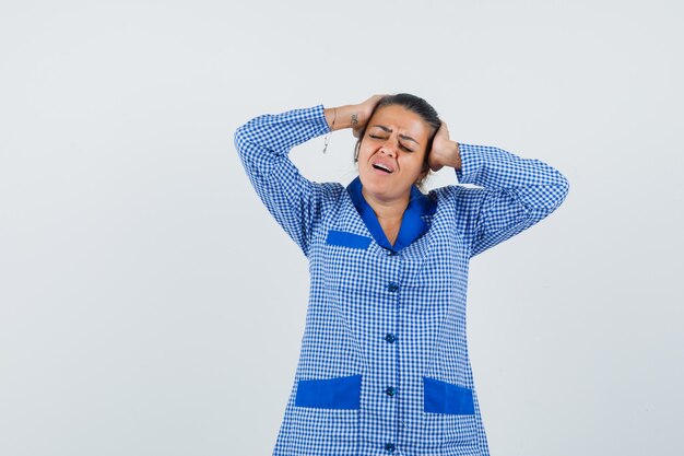 Mujer joven en camisa de pijama de cuadros vichy azul poniendo la mano en la cabeza, con dolor de cabeza y mirando molesto, vista frontal.