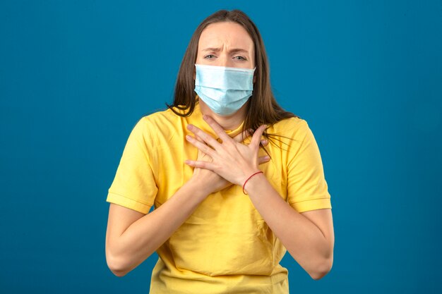 Mujer joven en camisa polo amarilla y máscara protectora médica que parece enferma con dolor en el pecho de pie sobre fondo azul.