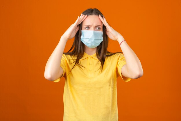 Mujer joven en camisa polo amarilla y máscara protectora médica tocando la cabeza sintiendo dolor de cabeza sobre fondo naranja aislado