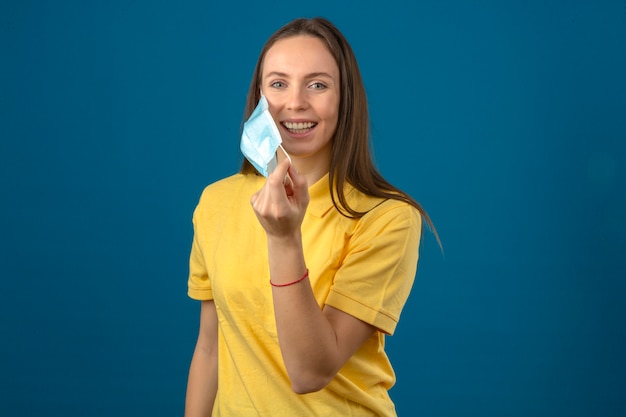 Mujer joven en camisa polo amarilla quitándose la máscara protectora médica sonriendo con cara feliz aislado sobre fondo azul.