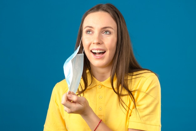 Mujer joven en camisa polo amarilla quitándose la máscara protectora médica sonriendo con cara feliz aislado sobre fondo azul.