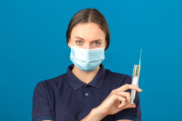 Mujer joven con camisa polo azul en máscara médica protectora con jeringa mirando a la cámara con cara seria de pie sobre fondo azul aislado