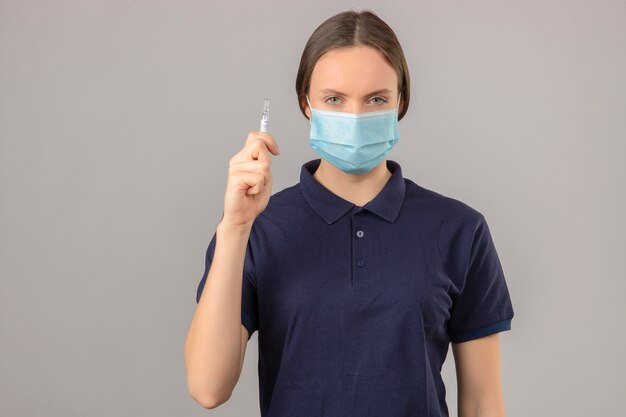 Mujer joven con camisa polo azul en máscara médica protectora sosteniendo una ampolla con una vacuna mirando a la cámara con cara seria de pie sobre fondo gris aislado