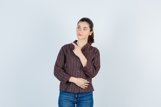 Mujer joven en camisa a rayas, jeans apuntando hacia la izquierda con el dedo índice y mirando enfocado, vista frontal.