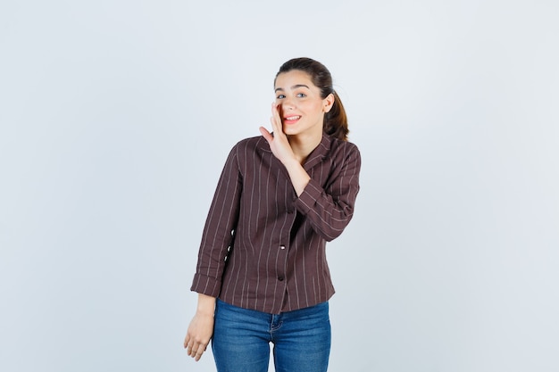 Mujer joven en camisa a rayas, jeans con la mano cerca de la boca como diciendo un secreto y mirando feliz, vista frontal.