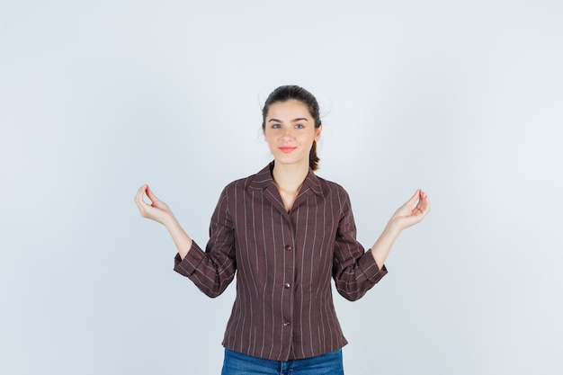 Mujer joven en camisa a rayas, jeans de pie en pose de meditación y mirando alegre, vista frontal.