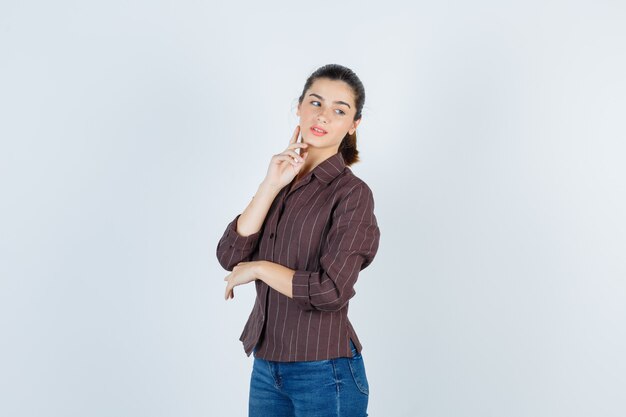 Mujer joven en camisa a rayas, jeans poniendo el dedo índice en la papada, mirando a otro lado mientras posa y luce encantador, vista frontal.