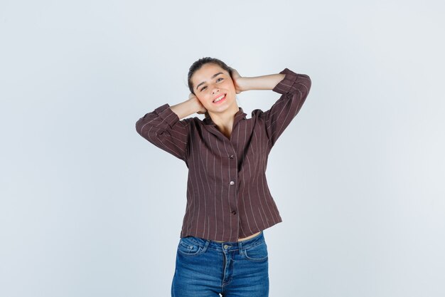 Mujer joven en camisa a rayas, jeans presionando las manos en las orejas y mirando alegre, vista frontal.