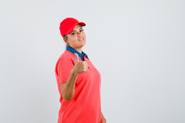 Mujer joven con camisa roja y gorra mostrando el pulgar hacia arriba y mirando bonita vista frontal.