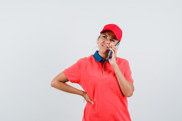 Mujer joven con camisa roja y gorra sosteniendo la mano en la cintura mientras habla por teléfono y mira pensativa, vista frontal.