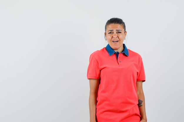 Mujer joven en camisa roja de pie derecho, haciendo muecas y posando mirando molesto, vista frontal.