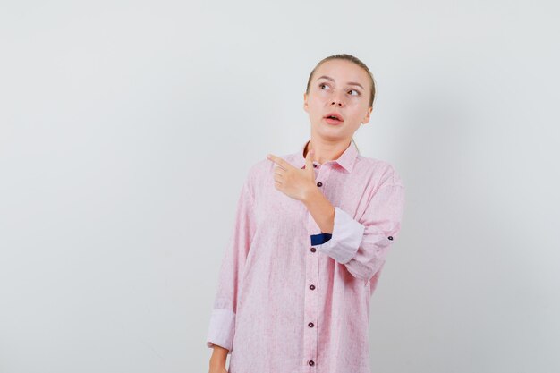 Mujer joven en camisa rosa apuntando hacia afuera y mirando enfocado